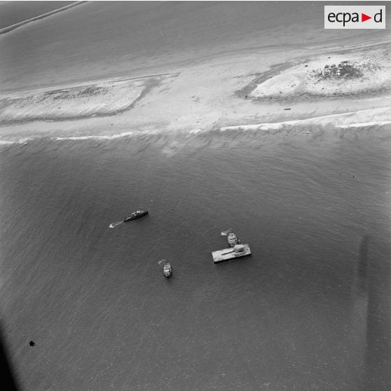 Vue aérienne du site du chantier, de bâtiments pousseurs et d'une barge, et du camp de tentes sur un motu. Construction du site de Moruroa.