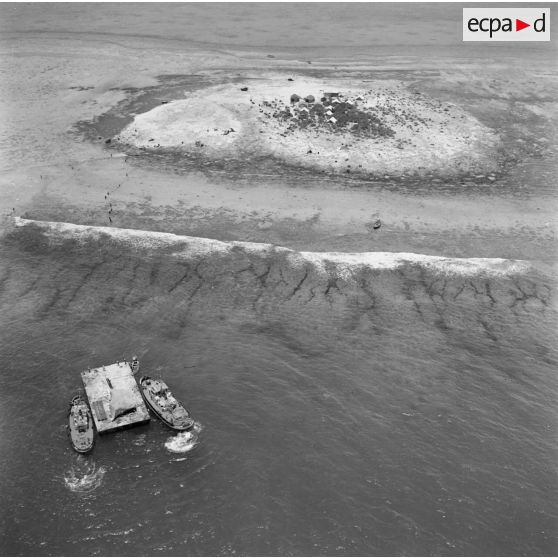 Vue aérienne du site du chantier, de bâtiments pousseurs et d'une barge, et du camp de tentes sur un motu. Construction du site de Moruroa.