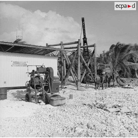 Personnels du génie militaire, shelter sous abri, bétonnière et camion grue. Peut-être sur l'atoll d'Hao.