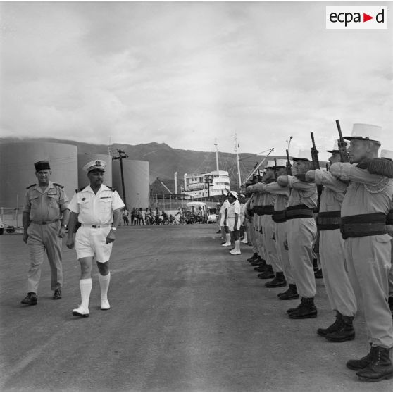 Revue des troupes, dont des légionnaires, par le capitaine de vaisseau Jean Pétesch, lors de la fin de son commandement du Centre d'expérimentations du Pacifique (CEP), en compagnie d'un commandant, port militaire de Fare ute à Papeete, mars 1964. En arrière-plan, le pétrolier tanker M.E. Lombardi.