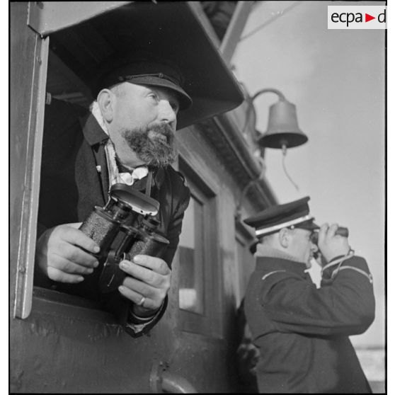 Le patron d'un chalutier armé par la Marine nationale et un enseigne de vaisseau observent l'horizon aux jumelles depuis le poste de navigation.