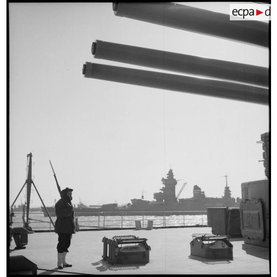 Sentinelle en faction sous une des tourelles du cuirassé Dunkerque. A  l'arrière-plan, le cuirassé Strasbourg amarré.