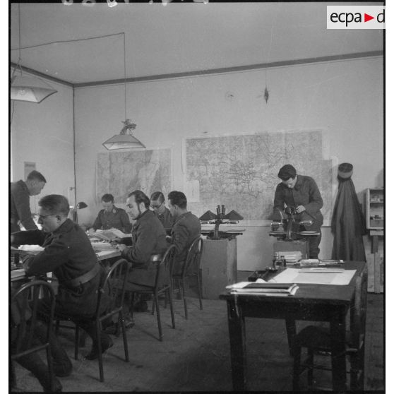 Plan général d'une salle d'état-major où divers officiers travaillent sur des photographies aériennes.