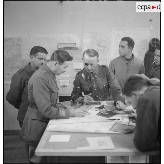 Plan général d'une salle d'état-major où divers officiers travaillent sur des photographies aériennes.