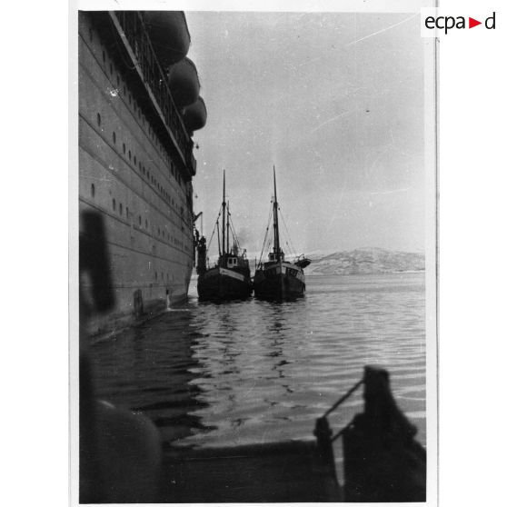 Deux bateaux de pêche accostent le flanc d'un paquebot /transport de troupes dans un fjord.