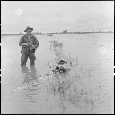 Un parachutiste armé d'un pistolet-mitrailleur MAT 49 se tient à côté du cadavre d'un partisan Viêt-minh flottant dans l'eau de la rizière.