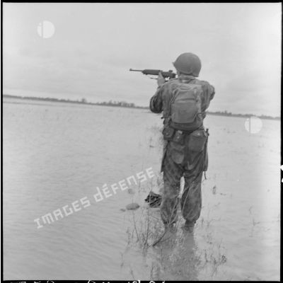 Un parachutiste visant avec sa carabine US M1 se tient dans une rizière inondée.  A ses pieds flotte le cadavre d'un partisan Viêt-minh.