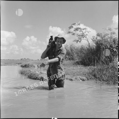 Le cinéaste Pierre Schoendoerffer progresse dans une rizière avec une caméra Bell&Howell sur l'épaule au cours de l'opération Claude dans le secteur de Tien Lang.