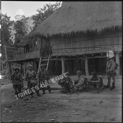 Arrivé en fin de journée au village laotien de Ban Loi, un groupe de parachutistes du 5e BPVN de la colonne Langlais pose avec son armement devant une maison traditionnelle sur pilotis au toit de chaume.