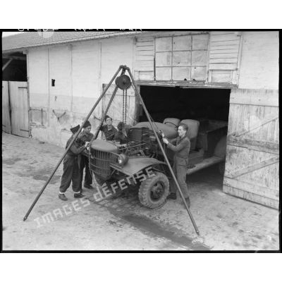 Des apprentis manipulent un lève-moteur au dessus d'un véhicule militaire, à l'entrée d'un atelier.