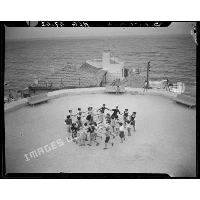 Plan en plongée d'enfants qui font une ronde dans une cour qui surplombe la mer, de la maison de l'enfance de la division d'Alger, à Raïsville.