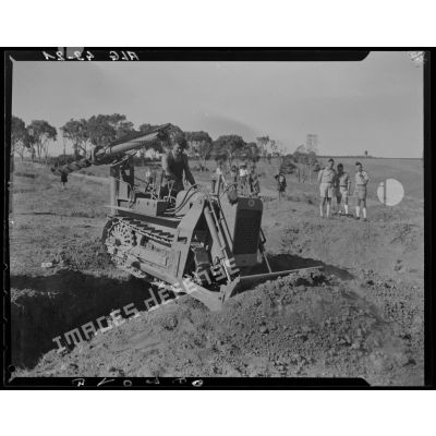 Un militaire nivèle un terrain avec un bulldozer équipé d'une tarière, sous le regard d'autorités militaires.