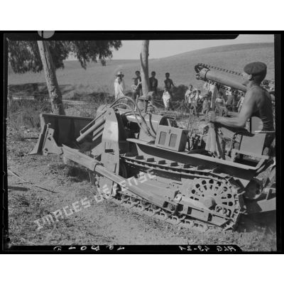 Un militaire déboise un terrain avec un bulldozer équipé d'une tarière, sous le regard de la population civile.
