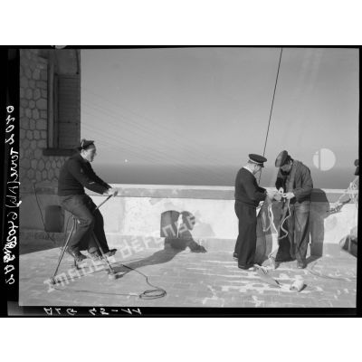 Trois marins identifient et réparent une panne de courant ; l'un d'eux sur un pédalier électrique, sur la terrasse d'un sémaphore, face à la mer.