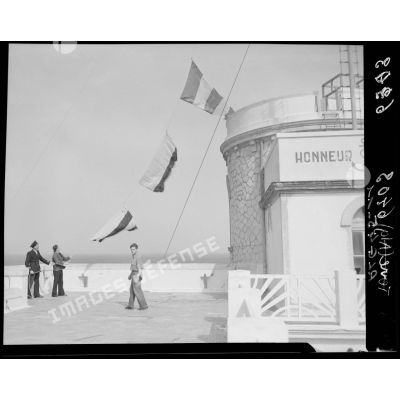 Trois marins, dont le chef de poste, effectuent la reconnaissance d'un bâtiment à l'aide d'un pavillon du code, sur la terrasse d'un sémaphore, face à la mer.