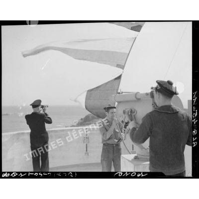 Trois marins, dont le chef de poste, effectuent une transmission par pavillon et projecteur, sur la terrasse d'un sémaphore, face à la mer.
