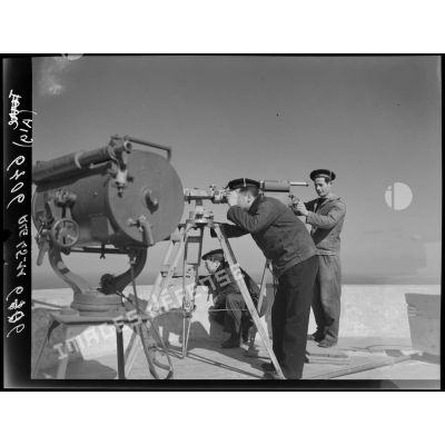 Trois marins interprètent des signaux d'un bâtiment, à l'aide d'un projecteur et de jumelles sur trepied, sur la terrasse d'un sémaphore, face à la mer.