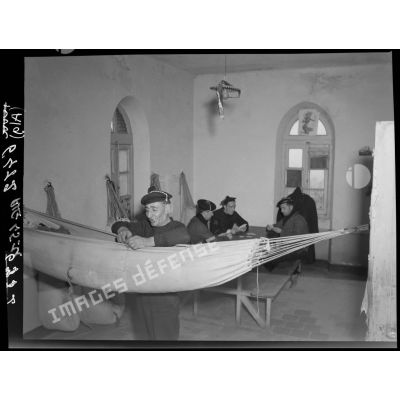 Un marin prépare son hamac, pendant que trois autres, en arrière plan, jouent aux cartes, dans le dortoir d'un sémaphore.