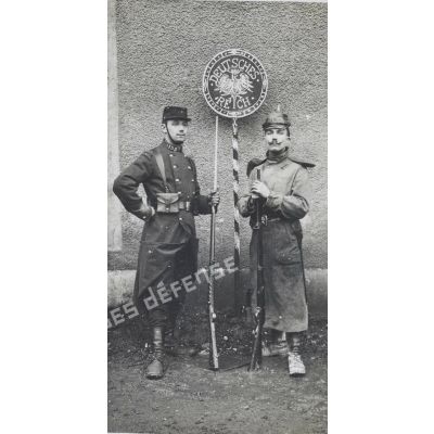 [Portrait en pied de deux soldats du 153e régiment d'infanterie, dont l'un porte un uniforme allemand approximatif, de part et d'autre d'un panneau de signalisation de la frontière avec le "Reich" allemand].