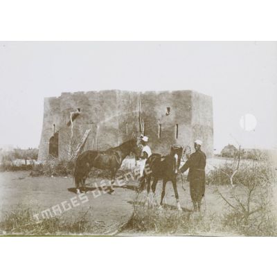 [Deux gardes africains et leurs chevaux devant un fort en terre crue.]