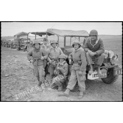 Vie quotidienne au 8e RIM. Portrait d'un groupe de soldats assis sur le devant d'une jeep. En arrière plan, un convoi militaire à l'arrêt. [légende d'origine]