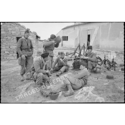 Vie quotidienne au 8e RIM. Portrait d'un groupe de soldats assis. Repos pendant une manoeuvre, les armes sont disposées en faisceau. En arrière plan, une bâtisse. [légende d'origine]
