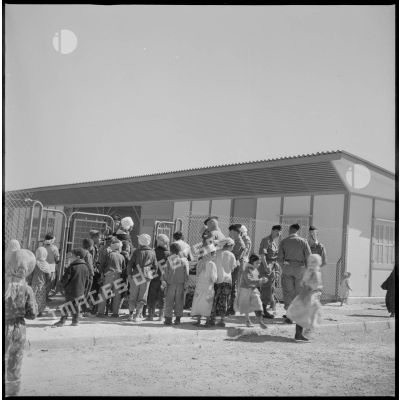 Devant un bâtiment, des soldats effectuent une distribution auprès des enfants de Wagram. [légende d'origine]