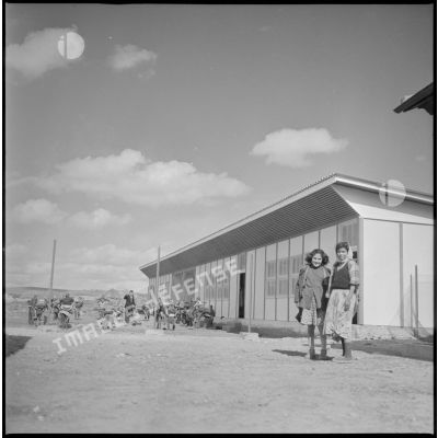 Tournée de secteur près de Tamesna et de Fenouane. Portrait de deux jeunes filles dans la cours de leur école. En arrière-plan, des salles de classes devant lesquelles des enfants jouent. [légende d'origine]
