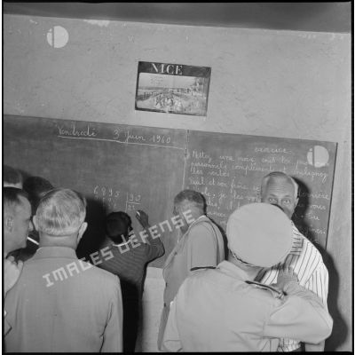 Les autorités civiles et militaires visitent une salle de classe. La date du vendredi 3 juin 1960 est inscrite sur le tableau de la classe. Au centre du tableau, un élève pose une division. [légende d'origine]
