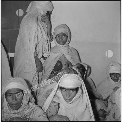 Portrait d'un groupe de femmes et d'enfants assis dans une salle d'attente. Un nourrisson tête au sein de sa mère. [légende d'origine]