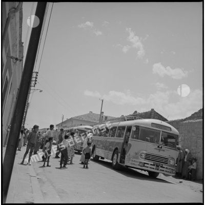En ville, deux bus venus récupérer des enfants stationnent en attendant le départ pour le Camp des chasseurs du 8e RIM. Plusieurs enfants accompagnés de leurs parents sont visibles devant les bus. [légende d'origine]