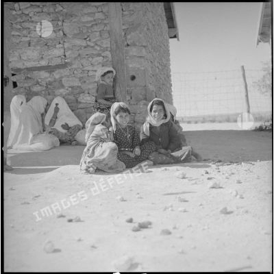 [Portrait d'enfants et de femmes assis au sol, dans un centre de regroupement. Les femmes, à l'ombre d'une bâtisse, se couvrent le visage.]