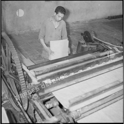 [Papeterie d'Aïn-el-Hadjar. Un ouvrier, avec des feuilles à la main, se tient devant une machine utilisée pour la fabrication du papier.]