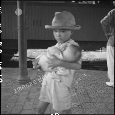 Sur une quai de la gare d'Hanoï, un petit garçon tient un nourrisson dans ses bras.