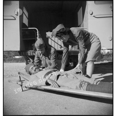 Un tirailleur blessé est installé sur un brancard avant d'être hissé à bord d'une ambulance par deux conductrices ambulancières du 27e escadron du Train.