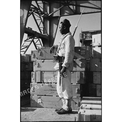 Portrait en pied d'un tirailleur sénégalais montant la garde sur le port de Casablanca auprès de caisses de matériel.