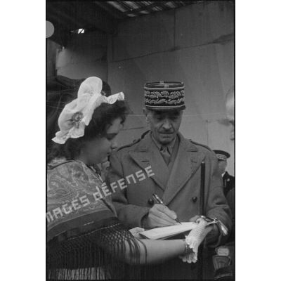 Après le discours du général de Gaulle, le général d'armée Catroux signe le livre d'or de la résistance que lui présente une jeune fille en costume traditionnel alsacien.