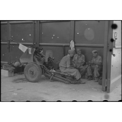 Sur le terrain d'aviation Naples-Pomigliano, trois parachutistes de la 1.Fallschirm-Jäger.Division mangent à l'ombre d'un hangar, l'un d'eux est assis sur un canon antiaérien de 2 cm FlaK 30 aérotransportable (Lufttransportfähige).