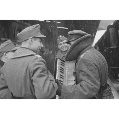 Sur le quai d'une gare, des permissionnaires allemands rejoignent leur affectation, ici un officier joue de l'accordéon.