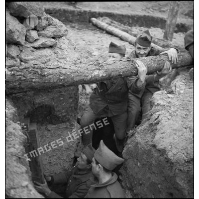 Photographie de groupe d'artilleurs du 185e RA qui déplacent un tronc de résineux afin de couvrir une tranchée.