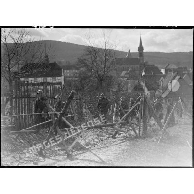 Un groupe de soldats de la 5e armée se déplace en colonne à la sortie d'un village dans un réseau de barbelés.
