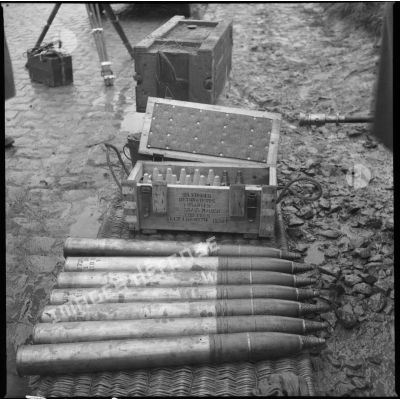 Plan moyen d'obus de 75 mm CA alignés au sol avec en arrière-plan une caisse de fusées.