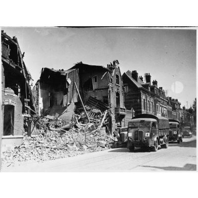 Des camions Renault AGR de la 1re DIM roule de trois quarts face en plan général dans une rue détruite de Valenciennes.
