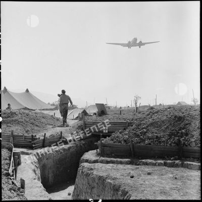 Un avion de transport Dakota survole le camp retranché de Diên Biên Phu dont on aperçoit l'entrée d'une antenne chirurgicale.