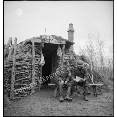 A l'entrée d'un abri fortifié de la 2e armée, deux soldats assis lisent un journal.