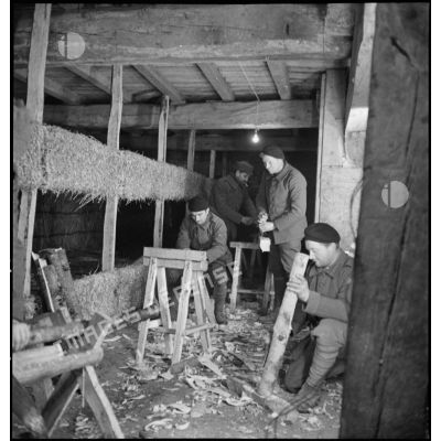 Des soldats de la 2e armée équarrissent des morceaux de bois à l'intérieur d'un abri de campagne garni de châlits.