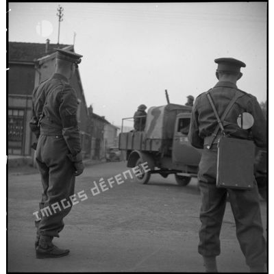 Deux soldats de la BEF, photographiés de dos dans un village, regardent passer un convoi.