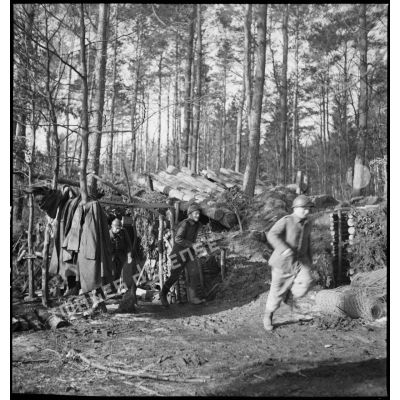 Dans un bois, des artilleurs de la 5e armée sortent en courant d'un abri de rondins.