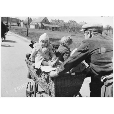 Sur une route, un homme pousse un landau contenant deux enfants.