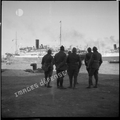 Sur le port de Marseille des soldats de la 6e armée, photographiés de dos, regardent un cargo à quai.